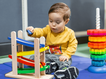 A child is interacting with colorful educational toys on a wooden table. The child’s face is obscured for privacy. On the left, there’s a wooden race track with red, yellow, green, and blue lanes where the child is placing a small blue car at the top. To the right of the race track stands a tall stacking toy with multiple colored rings in shades of purple, blue, green, yellow, orange, and red arranged in descending order from largest at the bottom to smallest at the top.